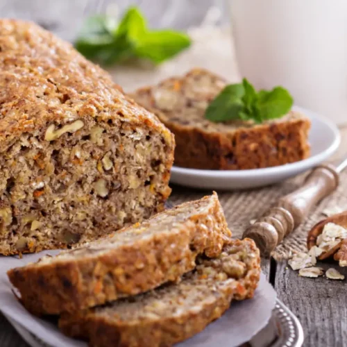 A loaf of banana bread with visible nuts and oats, sliced and displayed on a rustic wooden surface, with a cup of tea and fresh mint leaves in the background.