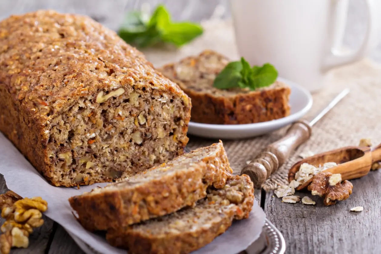 A loaf of banana bread with visible nuts and oats, sliced and displayed on a rustic wooden surface, with a cup of tea and fresh mint leaves in the background.