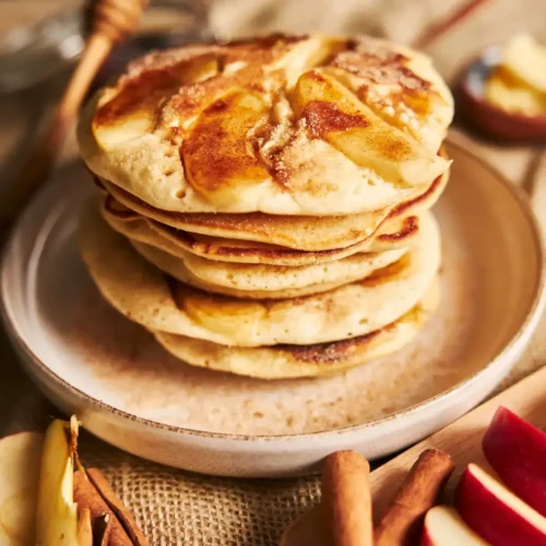 A close-up stack of apple pancakes topped with cinnamon and sugar, served on a ceramic plate with apple slices and cinnamon sticks nearby.