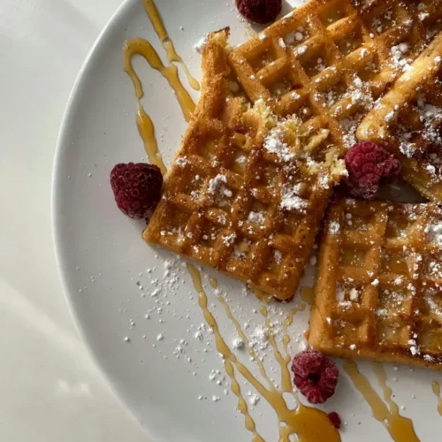 Golden homemade waffles drizzled with syrup and dusted with powdered sugar, garnished with fresh raspberries on a white plate.