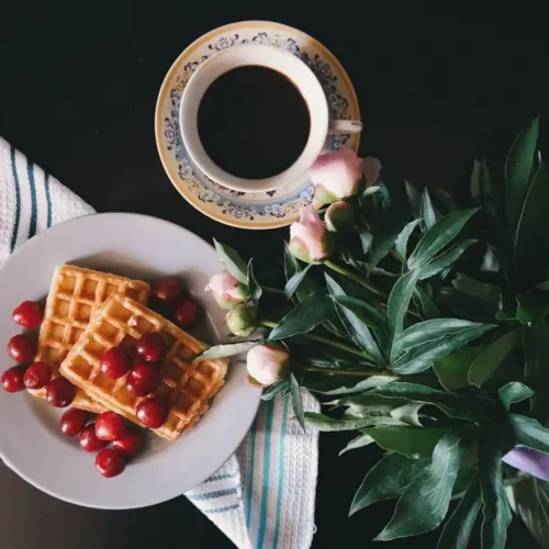 A cozy breakfast setup featuring two golden waffles topped with fresh cherries, accompanied by a cup of coffee and a bouquet of flowers.