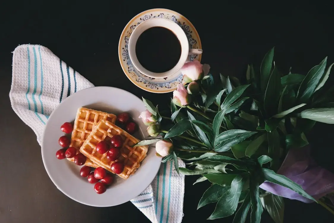 A cozy breakfast setup featuring two golden waffles topped with fresh cherries, accompanied by a cup of coffee and a bouquet of flowers.