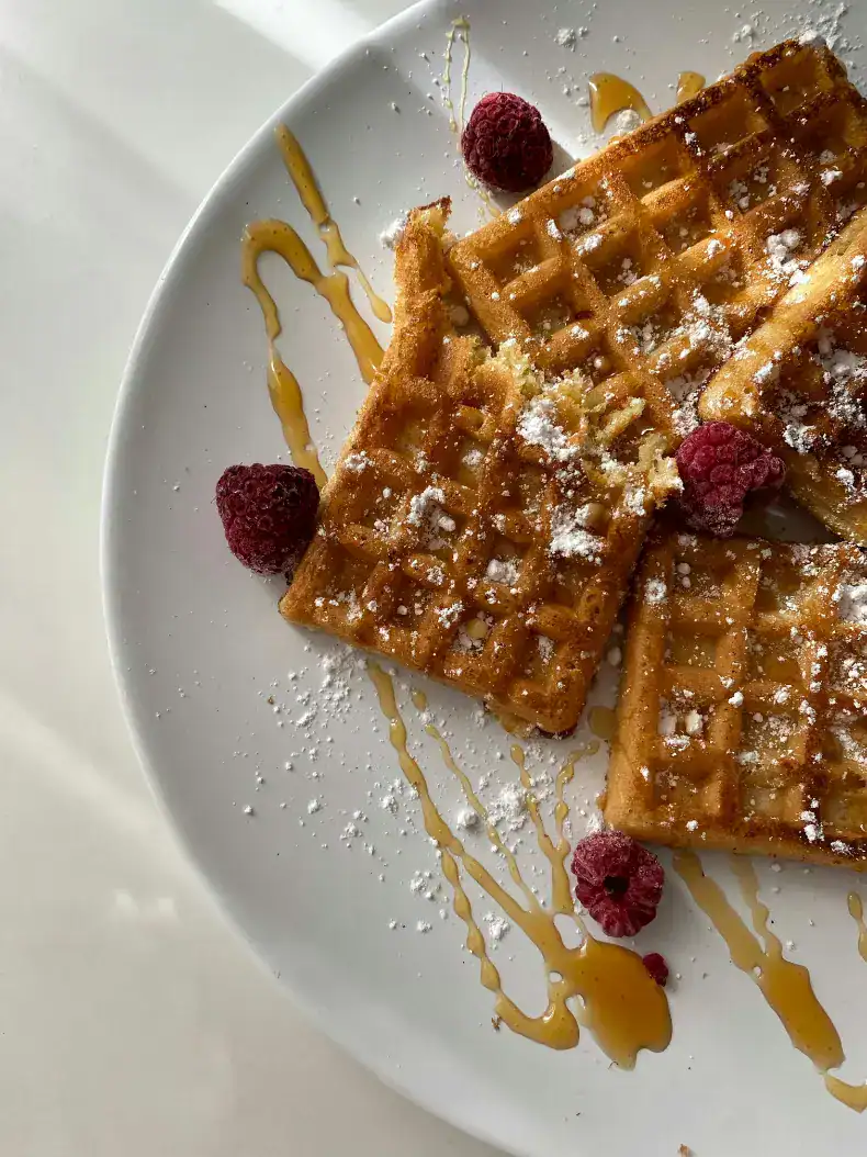 Golden homemade waffles drizzled with syrup and dusted with powdered sugar, garnished with fresh raspberries on a white plate.