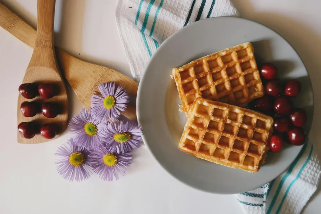 A plate with two golden waffles and fresh cherries, accompanied by wooden spoons holding cherries and purple flowers, creating a visually appealing breakfast scene