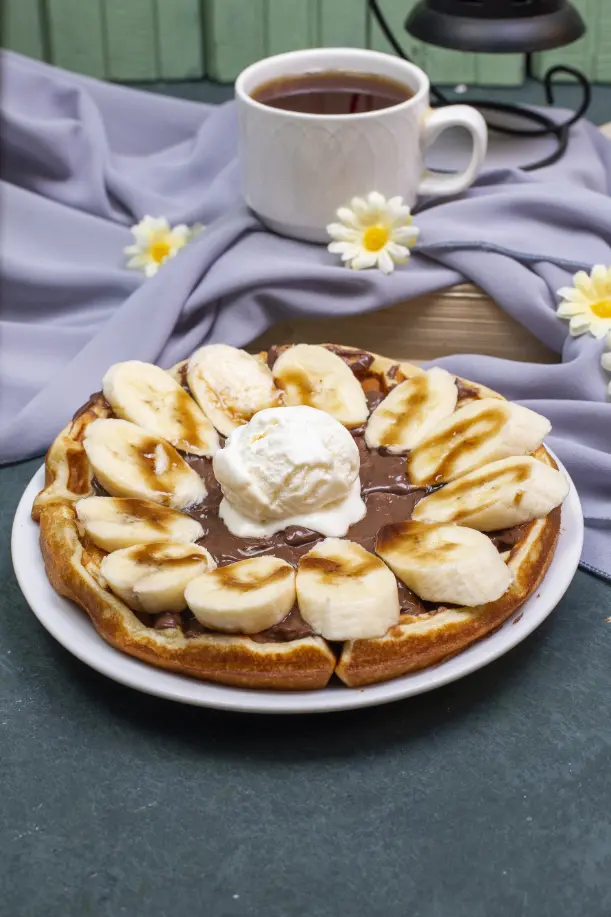 A plate of waffles topped with banana slices, caramel sauce, and a dollop of whipped cream, accompanied by a cup of tea on a purple cloth background.