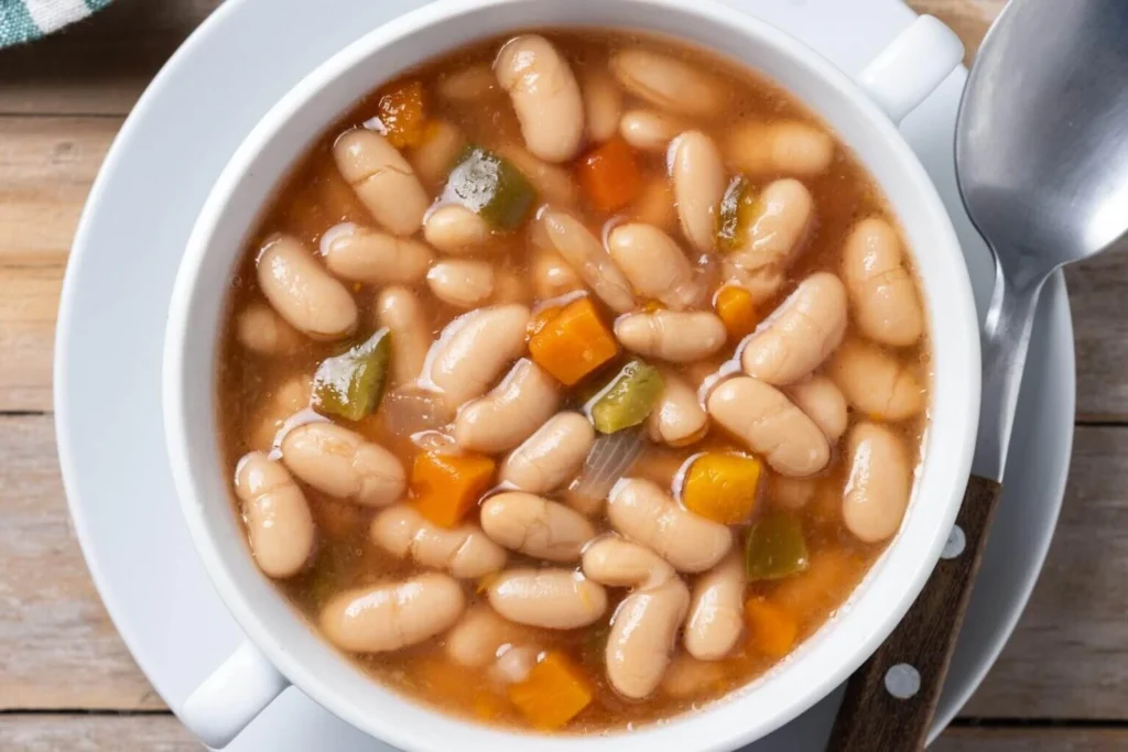 A hearty bowl of white bean stew with chunks of carrots and green bell peppers, served in a white bowl with a spoon on the side.