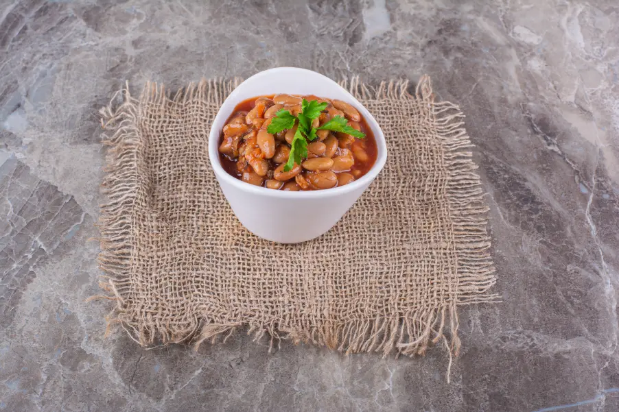 A white bowl of bean stew placed on a square burlap mat on a marble-textured surface, garnished with fresh parsley.