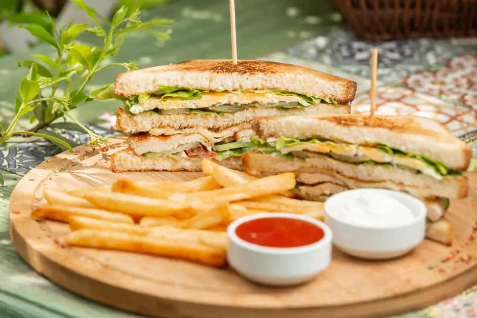 A wooden platter with two stacked club sandwiches filled with grilled chicken, crispy bacon, lettuce, cheese, and creamy sauce, served with golden French fries and dipping sauces.