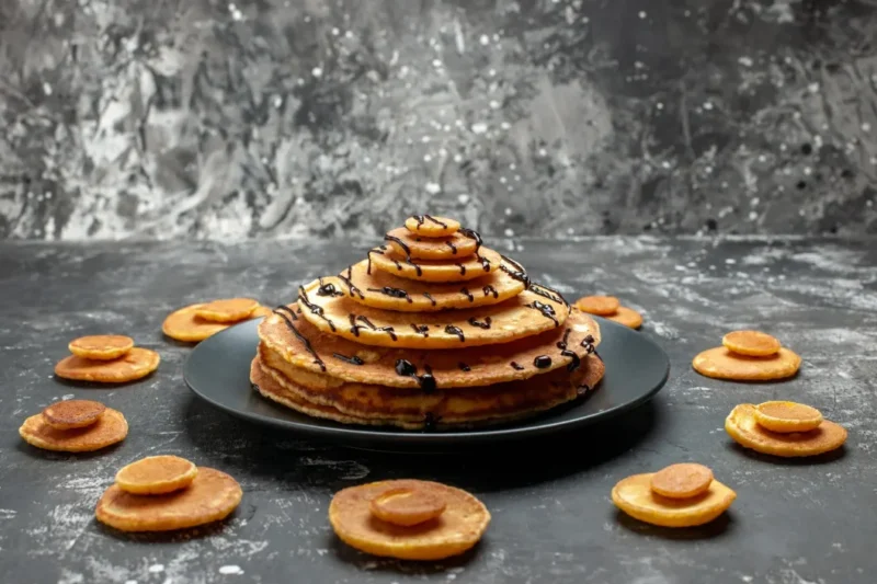 A creative stack of griddlecakes arranged in a pyramid shape, drizzled with chocolate syrup, and surrounded by mini pancake decorations on a dark, textured background.