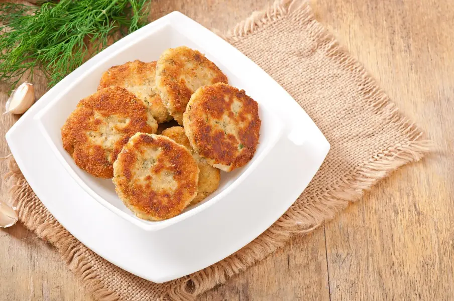 Golden, crispy chicken cutlet patties served on a white plate, placed on a rustic wooden table with fresh dill and garlic cloves nearby.