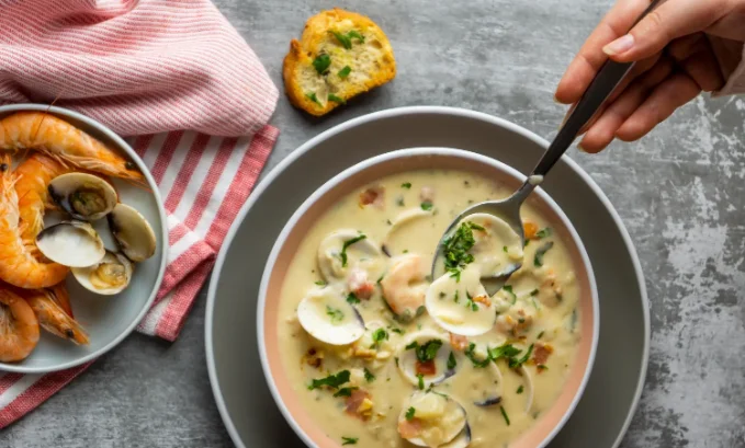 A creamy bowl of clam chowder filled with fresh clams, shrimp, and bacon, garnished with parsley. A person lifts a spoonful, while a plate of seafood and toasted bread sits nearby.