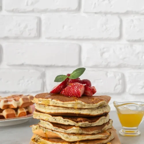 A stack of fluffy griddlecakes topped with fresh strawberries and mint, drizzled with honey, served on a wooden board with waffles and syrup in the background against a white brick wall.