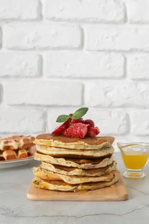 A stack of fluffy griddlecakes topped with fresh strawberries and mint, drizzled with honey, served on a wooden board with waffles and syrup in the background against a white brick wall.