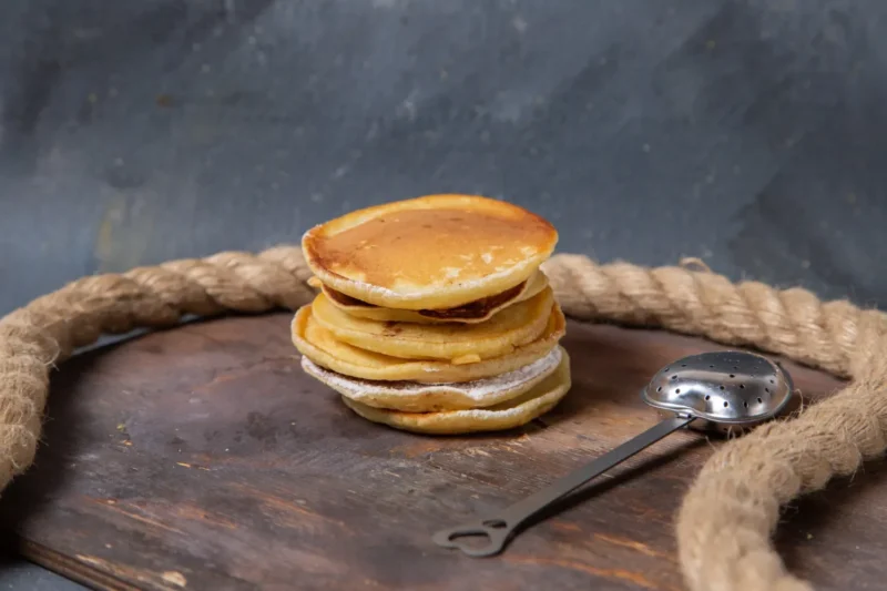 A stack of fluffy, golden-brown griddlecakes on a rustic wooden surface, with a metal tea infuser and thick rope in the background.