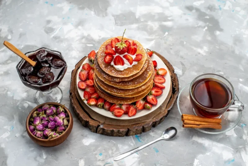 A tall stack of golden-brown griddlecakes topped with fresh strawberries, whipped cream, and powdered sugar, served with tea, cinnamon sticks, and strawberry jam on a rustic wooden platter.