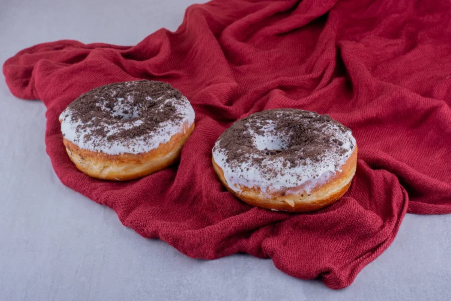 Two homemade doughnuts with white glaze and crushed chocolate cookies on a red fabric backdrop.