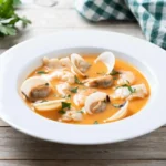 A bowl of fresh clam chowder with a rich, orange-hued broth, garnished with clams, fish, and fresh parsley, served on a rustic wooden table with a green checkered napkin.