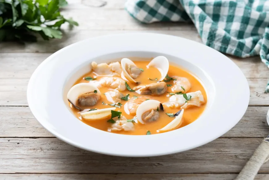 A bowl of fresh clam chowder with a rich, orange-hued broth, garnished with clams, fish, and fresh parsley, served on a rustic wooden table with a green checkered napkin.