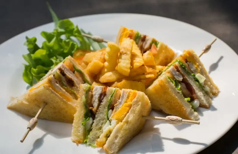 A plate of freshly made club sandwiches cut into quarters, filled with lettuce, tomato, cheese, and grilled chicken, served with crispy crinkle-cut fries and a side of green salad.