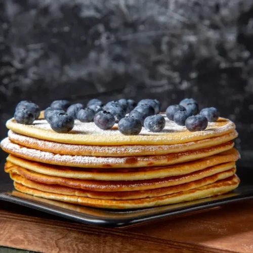 A stack of golden griddlecakes dusted with powdered sugar and topped with fresh blueberries, served on a black square plate against a dark textured background.