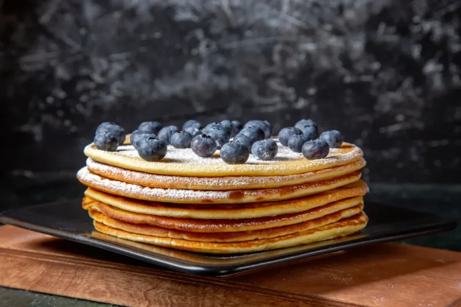 A stack of golden griddlecakes dusted with powdered sugar and topped with fresh blueberries, served on a black square plate against a dark textured background.