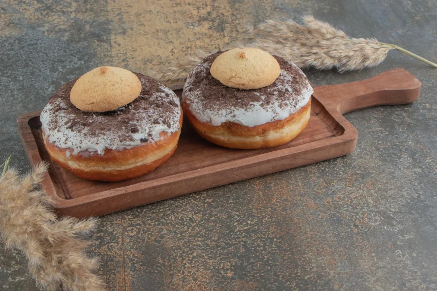 wo homemade no-yeast doughnuts with white glaze, crushed chocolate cookies, and a small cookie topping, served on a wooden board.