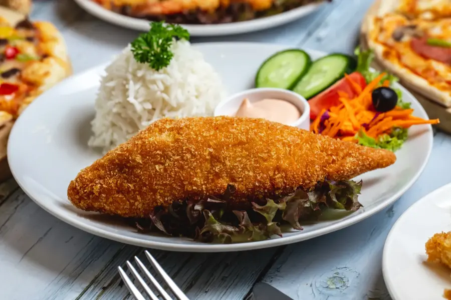 A crispy, golden-breaded chicken cutlet served with white rice, fresh salad, cucumber slices, and a dipping sauce on a white plate.