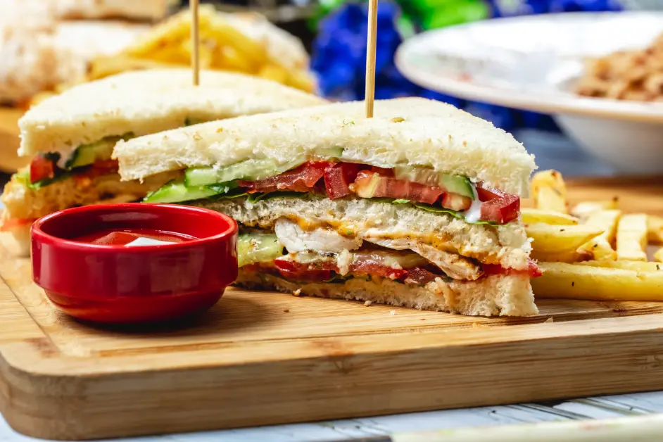 A fresh club sandwich with layers of grilled chicken, crispy bacon, lettuce, tomatoes, cucumbers, and creamy sauce, served with golden French fries and ketchup on a wooden board.