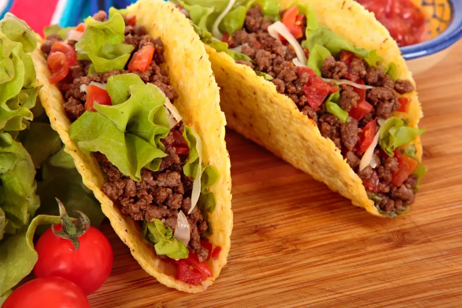 Two crispy hard-shell tacos filled with seasoned ground beef, fresh lettuce, diced tomatoes, and shredded cheese, served on a wooden board with cherry tomatoes and salsa on the side.