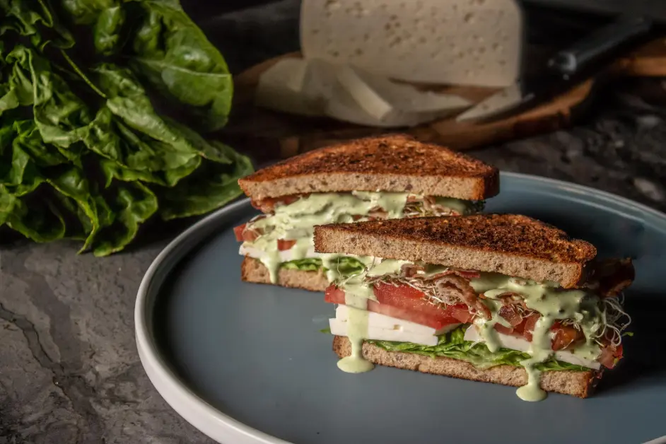 A gourmet club sandwich with toasted whole grain bread, fresh spinach, sliced tomatoes, creamy cheese, crispy bacon, and sprouts, drizzled with a green herb sauce, served on a blue plate.