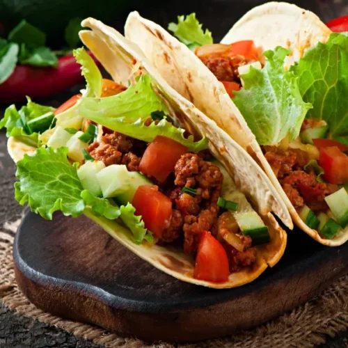 Two soft flour tortillas filled with seasoned shredded beef, fresh lettuce, diced tomatoes, and cucumbers, served on a rustic wooden board with a red chili pepper and fresh tomato in the background.