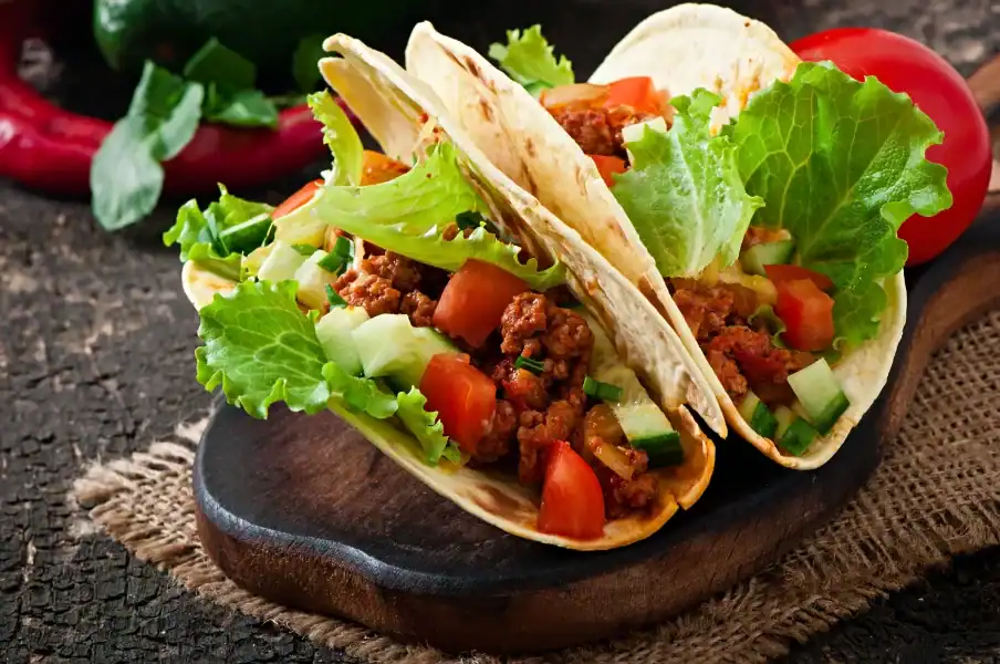 Two soft flour tortillas filled with seasoned shredded beef, fresh lettuce, diced tomatoes, and cucumbers, served on a rustic wooden board with a red chili pepper and fresh tomato in the background.