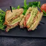 A fresh vegetarian club sandwich made with whole grain bread, avocado, tomatoes, lettuce, and a creamy spread, served on a dark slate plate with arugula and ripe tomatoes.