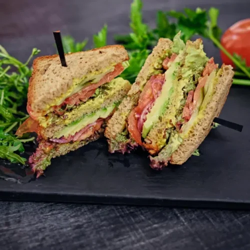 A fresh vegetarian club sandwich made with whole grain bread, avocado, tomatoes, lettuce, and a creamy spread, served on a dark slate plate with arugula and ripe tomatoes.