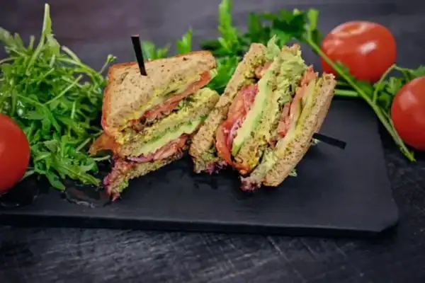 A fresh vegetarian club sandwich made with whole grain bread, avocado, tomatoes, lettuce, and a creamy spread, served on a dark slate plate with arugula and ripe tomatoes.