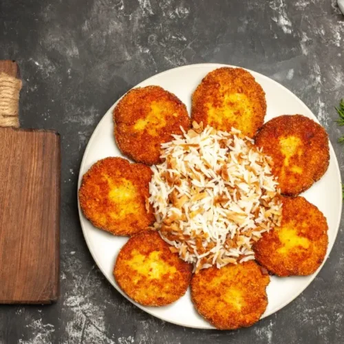 A plate of golden, crispy chicken cutlets arranged in a circular pattern around a mound of seasoned rice, with fresh tomatoes, dill, and garlic on the side.