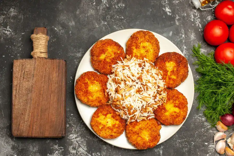 A plate of golden, crispy chicken cutlets arranged in a circular pattern around a mound of seasoned rice, with fresh tomatoes, dill, and garlic on the side.