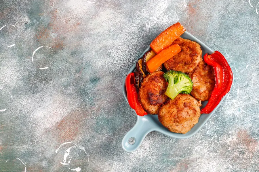 A serving of golden chicken cutlet patties with roasted carrots, red bell peppers, broccoli, and eggplant in a blue dish on a textured background.