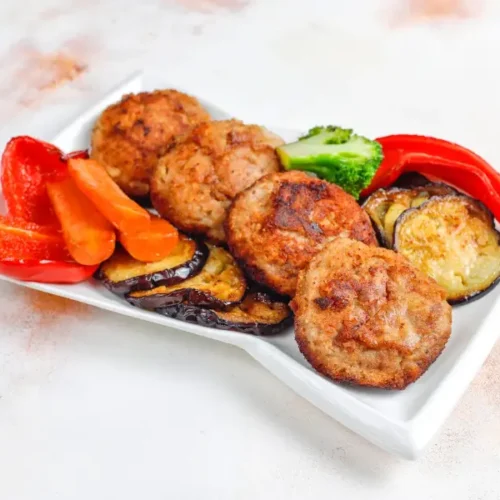 A plate of crispy chicken cutlet patties served with roasted eggplant, carrots, red bell peppers, and broccoli on a white dish.