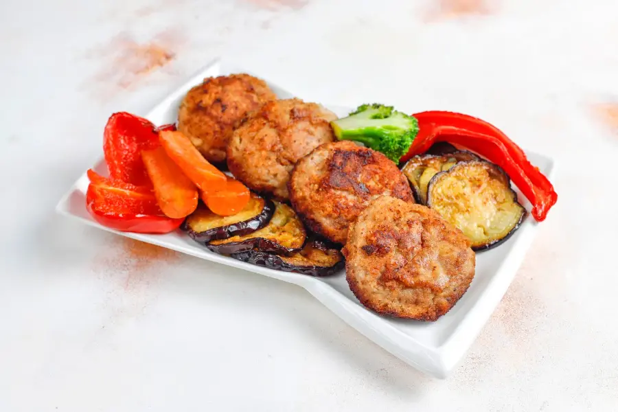 A plate of crispy chicken cutlet patties served with roasted eggplant, carrots, red bell peppers, and broccoli on a white dish.