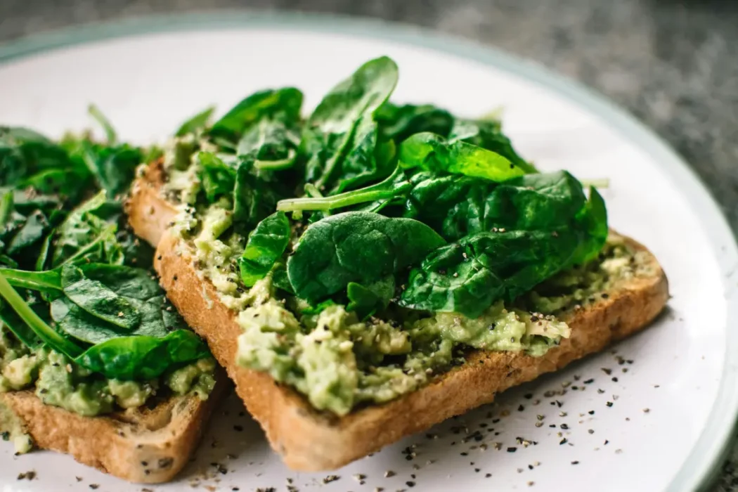 Two slices of whole grain toast topped with mashed avocado and fresh spinach, seasoned with black pepper, served on a white plate.