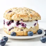 A freshly baked blueberry scone filled with whipped cream and blueberry jam, served on a white plate with fresh blueberries scattered around.