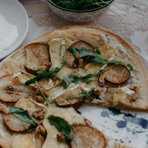 A gourmet fruit pizza topped with thinly sliced pears, melted cheese, walnuts, and fresh arugula, served on a rustic plate.