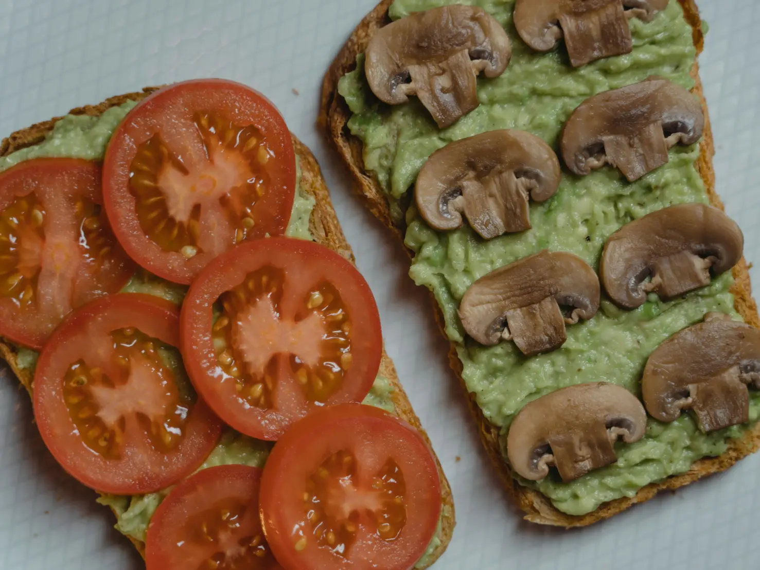 Two slices of whole grain toast topped with mashed avocado, one garnished with fresh tomato slices and the other with sliced mushrooms.