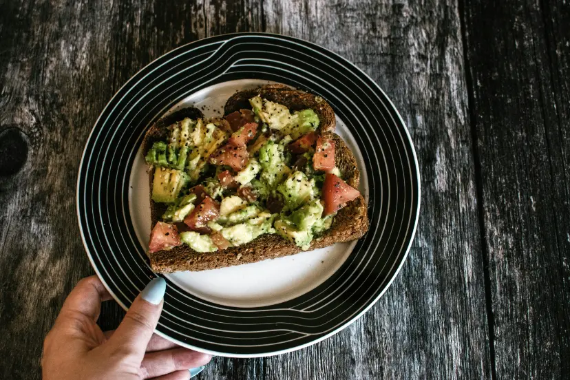 A slice of whole grain toast topped with chunky avocado, diced tomatoes, and a sprinkle of sesame seeds, served on a black and white plate.