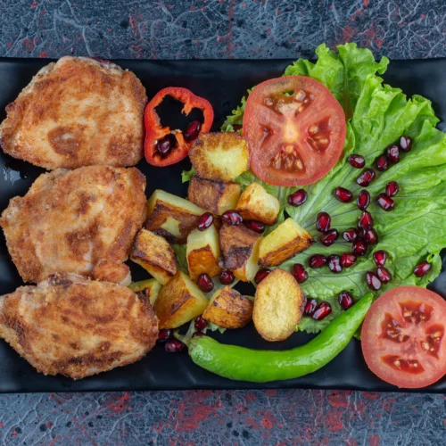 A black plate with crispy chicken cutlets, roasted potatoes, lettuce, sliced tomatoes, a green chili, pomegranate seeds, and grilled red bell pepper slices.