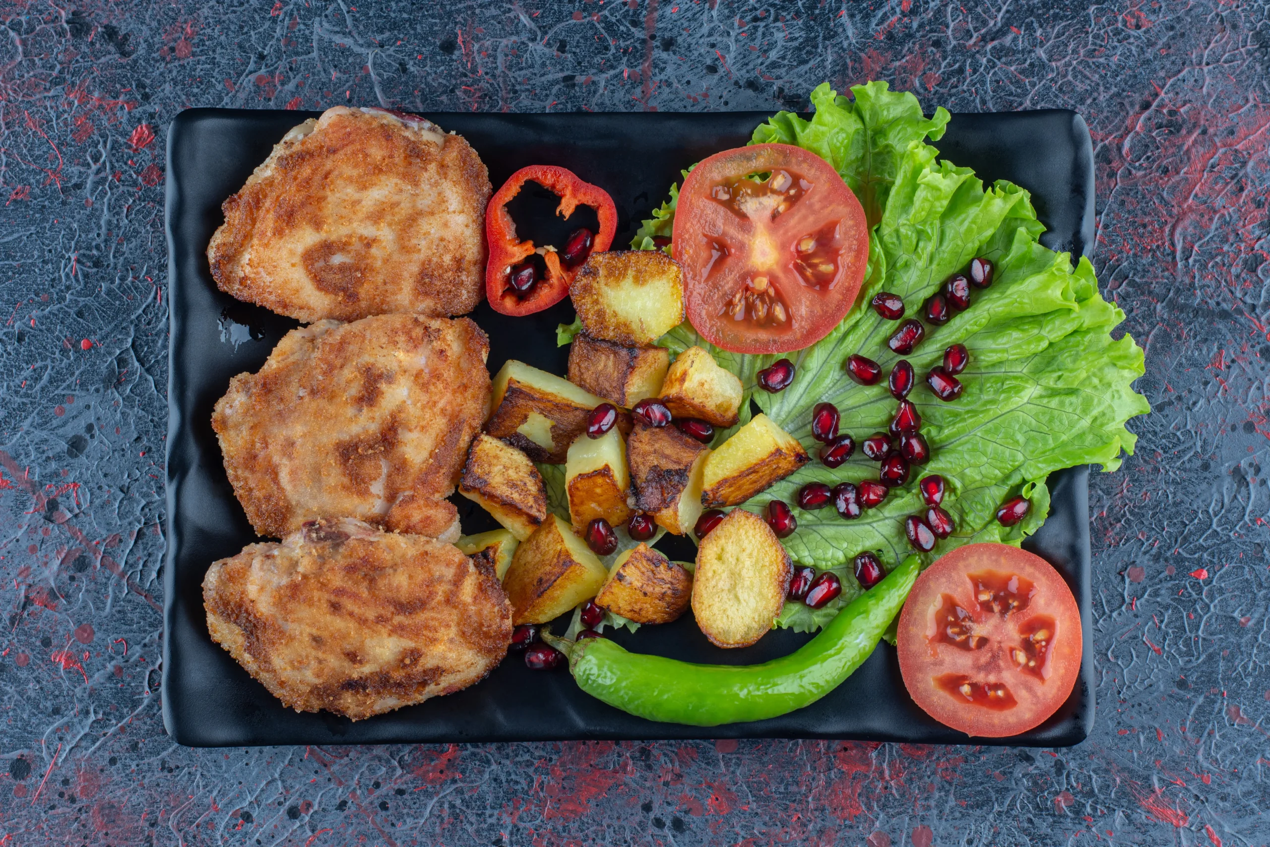 A black plate with crispy chicken cutlets, roasted potatoes, lettuce, sliced tomatoes, a green chili, pomegranate seeds, and grilled red bell pepper slices.