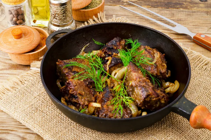 A cast iron skillet filled with perfectly cooked beef liver and caramelized onions, garnished with fresh dill, placed on a rustic wooden table.