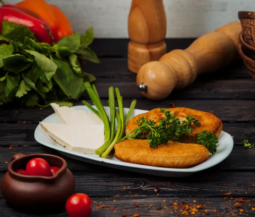 A plate with golden chicken cutlets, fresh parsley, green onions, and slices of white cheese, surrounded by ingredients like cherry tomatoes, herbs, and peppers on a wooden table.