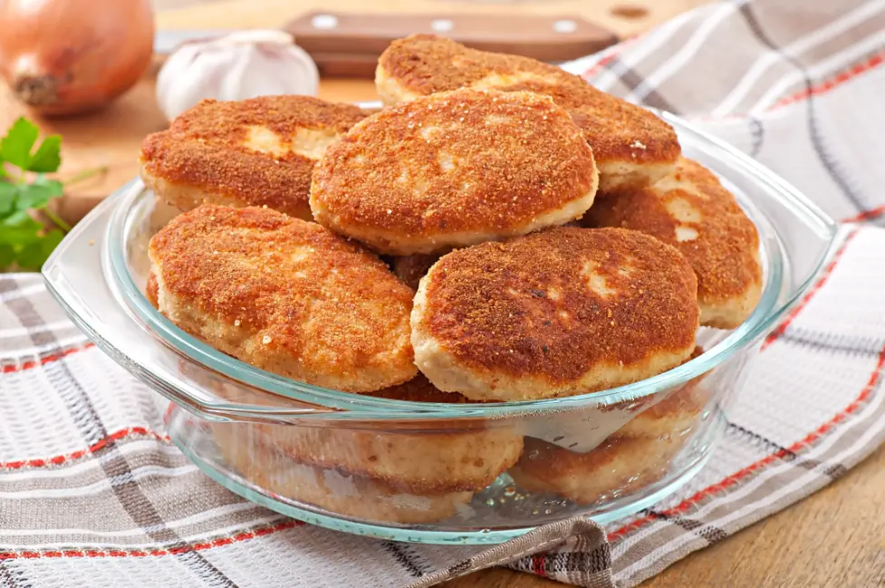 Golden-brown chicken cutlets stacked in a clear glass bowl, with onions, garlic, and parsley in the background.